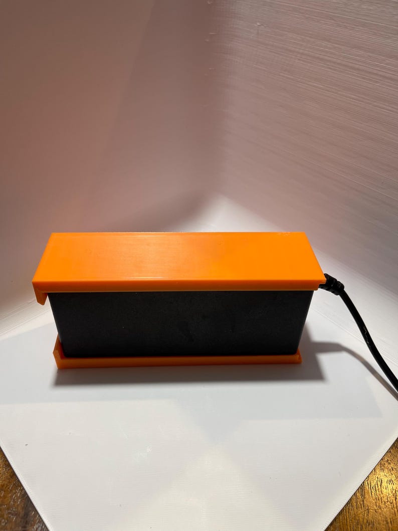 May include: A black rectangular box with an orange lid and a black cord attached to the side.