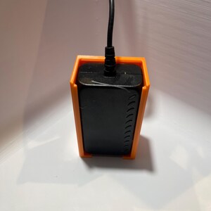 May include: A black power adapter with a black cord is held in place by an orange plastic wall mount.