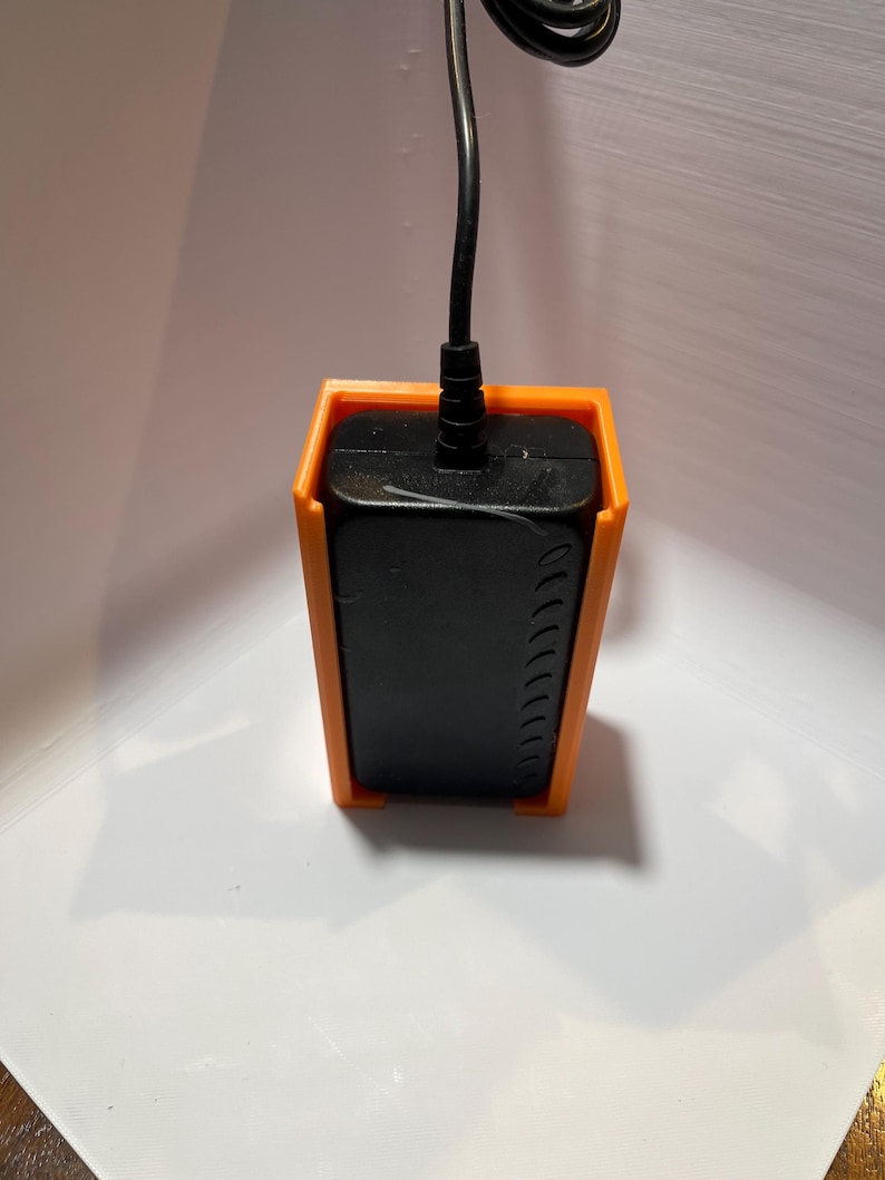 May include: A black power adapter with a black cord is held in place by an orange plastic wall mount.