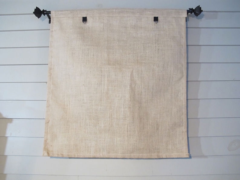 Puede incluir: Un tel&oacute;n de fondo de tela de arpillera de color blanco roto liso, colgado de una varilla de metal negro con terminales decorativos. El tel&oacute;n de fondo est&aacute; sujeto a la varilla con anillos de metal negro.
