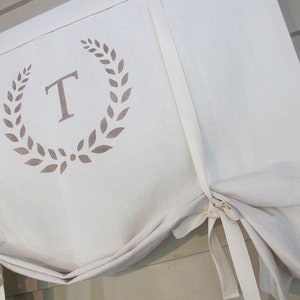 May include: White fabric window valance with a brown monogram of the letter "T" inside a wreath of leaves.