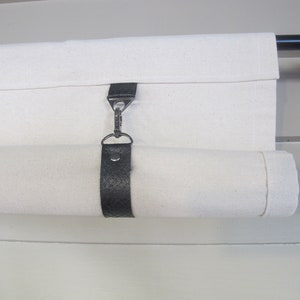May include: A white canvas shade with a black leather strap and a silver hook. The shade is rolled up and attached to a black metal rod.
