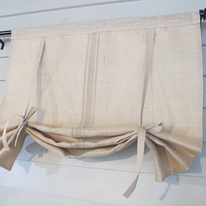 May include: A beige linen Roman shade with a rod pocket and tie-up ribbons. The shade has two vertical stripes and is mounted on a black metal rod with decorative finials. The fabric appears to be a natural fiber.