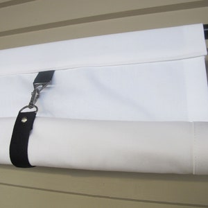 May include: White canvas shade rolled up on a black metal rod with a black strap and silver hook. The shade is mounted to a wall.