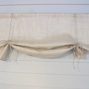 May include: A natural-colored burlap valance with a gathered, ruffled design. The valance is hung on a black metal rod with decorative finials.