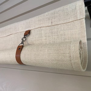 May include: A beige burlap curtain with a brown leather strap and a silver hook. The curtain is rolled up and hanging from a black metal rod.