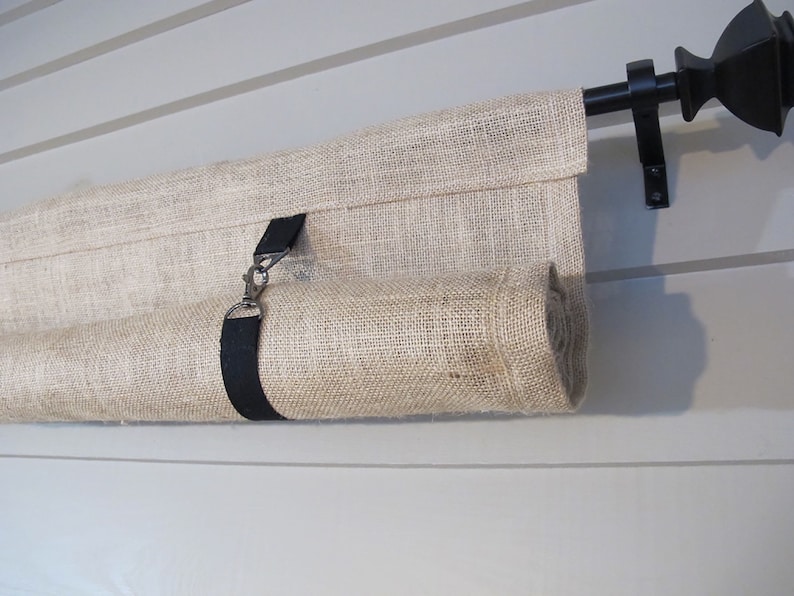 Puede incluir: Una cortina de arpillera beige enrollada y asegurada con una correa negra y un gancho plateado. La cortina est&aacute; montada en una varilla de metal negro con un remate negro.