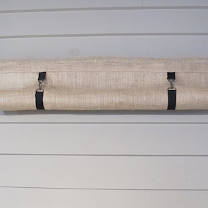 Puede incluir: Una cortina enrollable de lona beige con correas negras y clips plateados est&aacute; montada en una varilla de metal negro. La cortina est&aacute; enrollada y colgando de la varilla.
