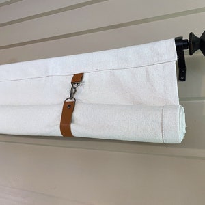 May include: A white canvas shade with a brown leather strap and a silver buckle is rolled up on a black metal rod with a decorative finial.