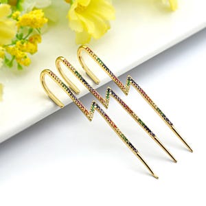 May include: Gold-toned lightning bolt earrings with a rainbow of colorful gemstones. The earrings have a curved hook design and a pointed bottom. The earrings are displayed on a white surface with yellow flowers in the background.