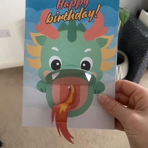 Könnte beinhalten: Eine Pop-up-Karte mit einem grünen Drachen, dessen rote und gelbe Zunge herausragt. Die Karte lautet "Happy birthday!" in blauem und weißem Text.