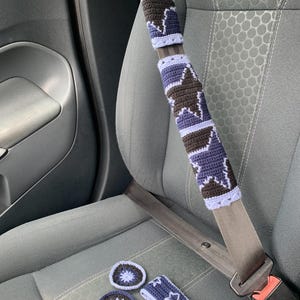 Op de afbeelding: Een gehaakte autogordelhoes met een herhalend sterpatroon in donkerbruin, marineblauw en lichtpaars. Inbegrepen zijn twee ronde gehaakte onderzetters, een klein rechthoekig gehaakt bandje en een kleine stervormige gehaakte hanger.