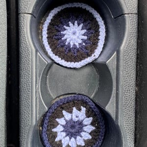 May include: Two circular crochet coasters featuring a starburst pattern in shades of light blue, dark blue, and black. Each coaster has a solid colored border and a central radiating design, crafted with yarn in a flat, round, and textured construction.