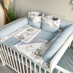 May include: A white crib with blue and white bedding featuring a whimsical elephant and rainbow design. The crib includes a blue waffle-weave bumper pad and two decorative baskets with elephant motifs.