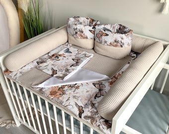 May include: A white cot with a beige and floral patterned cot sheet and bumper pads. There are two beige storage baskets with floral patterns and butterflies on the cot.