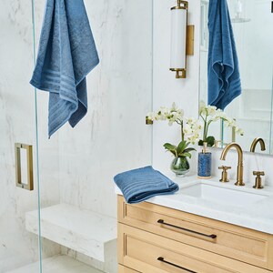 May include: A bathroom scene featuring blue towels, a light wood vanity with black hardware, and gold fixtures. A shower with a glass door and a marble bench is visible. Decorative elements include a vase of white orchids and a blue patterned soap dispenser.