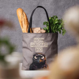 Può includere: Una borsa tote grigia con manici neri, caratterizzata da un'illustrazione di un uccello imbronciato e dalla scritta "HOW ABOUT NO" in caratteri stampatello. La borsa contiene una baguette lunga e un mazzetto di prezzemolo fresco.