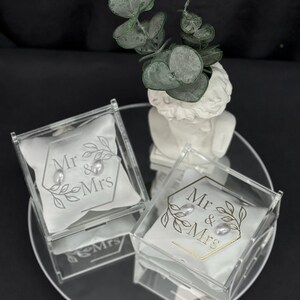 May include: Two clear acrylic ring boxes with white satin pillows, each holding a pearl. The pillows are printed with "Mr & Mrs" and a leaf design. The boxes sit on a mirrored surface, with a small white bust and greenery in the background.