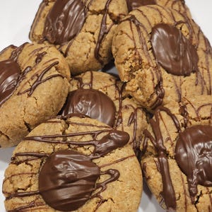 May include: A pile of freshly baked cookies. The cookies are round, with a light brown color and a textured surface. Each cookie is topped with a dark chocolate disc and drizzled with melted chocolate. A delicious treat.