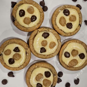 Peut inclure: Six biscuits ronds avec une cro&ucirc;te brun clair et une garniture jaune clair. La garniture est recouverte de trois p&eacute;pites de chocolat et de trois petits morceaux de p&acirc;te. Les biscuits sont sur une surface blanche avec des p&eacute;pites de chocolat &eacute;parpill&eacute;es autour.