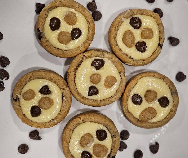 Peut inclure: Six biscuits ronds avec une cro&ucirc;te brun clair et une garniture jaune clair. La garniture est recouverte de trois p&eacute;pites de chocolat et de trois petits morceaux de p&acirc;te. Les biscuits sont sur une surface blanche avec des p&eacute;pites de chocolat &eacute;parpill&eacute;es autour.