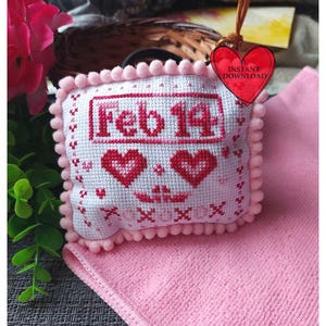 May include: A white cross-stitch pillow with pink pom-poms around the edge. The pillow features the text "Feb 14" in red and two red hearts. The pillow is sitting on a pink surface.