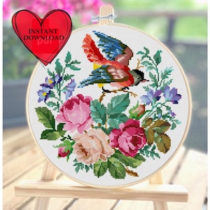 May include: A cross-stitch embroidery hoop featuring a colorful bird with red, blue, and yellow plumage, perched amidst a floral arrangement of pink and purple roses and greenery. The text "INSTANT DOWNLOAD" is in a red heart shape.