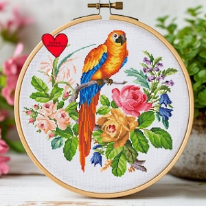 Könnte beinhalten: Ein Kreuzstich-Stickrahmen mit einem bunten Papagei auf einem Ast, umgeben von Blumen. Der Papagei hat orange, blaue und gelbe Federn. Die Blumen umfassen rosa und gelbe Rosen und Glockenblumen. Der Text "INSTANT DOWNLOAD" befindet sich auf einem roten Herz.
