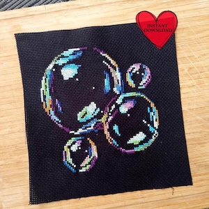 May include: A cross-stitch pattern of iridescent soap bubbles on a black background. The bubbles are various sizes and are overlapping. The pattern is framed and displayed on a wooden surface.