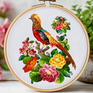 Może przedstawiać: Haft krzyżykowy przedstawiający kolorowego ptaka siedzącego na gałęzi, otoczonego żywymi kwiatami w drewnianym tamborku. Ptak ma odcienie czerwieni, pomarańczy, żółci i błękitu. Kwiaty obejmują różowe, pomarańczowe i żółte kwiaty.