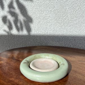 May include: A ceramic ashtray. The outer ring is light green, and the inner dish is light pink. It rests on a dark wooden surface. The background is a light grey wall with a shadow.