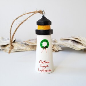 May include: A white wooden lighthouse ornament with a black and yellow striped roof. The lighthouse has a green wreath painted on the front and the words "Chatham Keegan Lighthouse" written below. The ornament has a brown twine string for hanging.