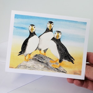 May include: A greeting card featuring a watercolor painting of three puffins. The birds have black and white plumage, orange feet, and distinctive beaks. They are standing on a rock with a blue and yellow background.