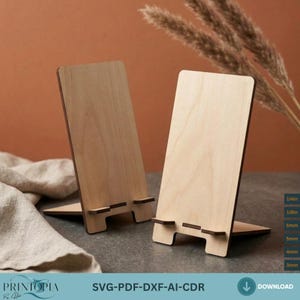 Pode incluir: Dois suportes de madeira clara para telemóvel com um painel traseiro retangular vertical e uma pequena saliência horizontal de apoio. Os suportes apresentam um design de encaixe para montagem e indicam espessuras de 6mm, 5mm, 4mm, 3mm.
