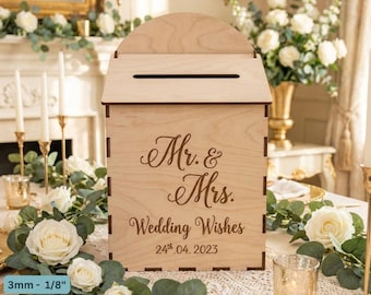 May include: A light-colored wooden wedding card box with a slot on top. The box is engraved with "Mr. & Mrs. Wedding Wishes 24th.04.2023". The box is surrounded by white roses and greenery.