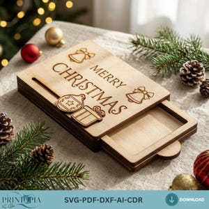 Könnte beinhalten: Eine rechteckige, helle Holzschachtel mit einem Schiebedeckel, der den eingravierten Text MERRY CHRISTMAS sowie zwei Glockensymbole, ein Christbaumkugelsymbol und ein Geschenksymbol zeigt. Der Deckel ist leicht geöffnet und gibt den Hohlraum frei.
