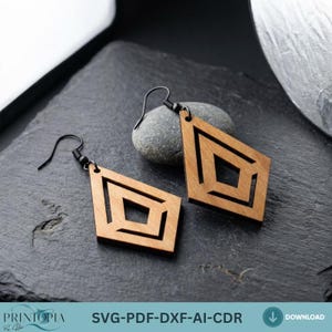 May include: Wooden geometric earrings with black hooks. The earrings feature a diamond shape with a square cut-out design. The earrings are light brown in color and are displayed on a dark gray surface.