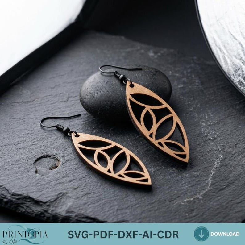 May include: A pair of wooden earrings with a leaf-like design. The earrings are light brown with a cutout pattern and black ear hooks. The earrings are displayed on a dark gray surface.