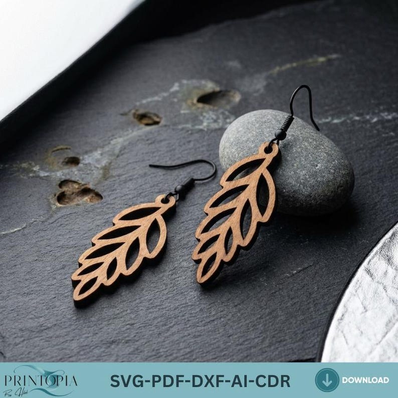 May include: A pair of wooden leaf earrings with black ear wires. The earrings are a light brown color and have a cutout leaf design. The earrings are displayed on a dark gray surface with a small gray stone.