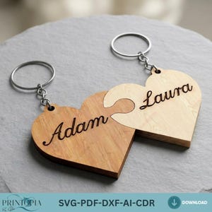 May include: Two wooden heart-shaped keyrings, designed to fit together like puzzle pieces. One keyring is engraved with "Adam," the other with "Laura." Each keyring has a silver ring attached. The background is a textured, grey surface.