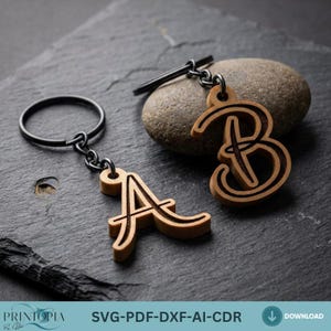 May include: Two wooden keychains, one with the letter "A" and the other with the letter "B". The keychains are light brown with a dark brown outline and are attached to a black metal ring and chain. The background is a dark gray stone surface.