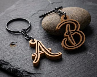 Puede incluir: Dos llaveros de madera, uno con la letra "A" y otro con la letra "B". Los llaveros son de color marrón claro con un contorno marrón oscuro y están unidos a un anillo y una cadena de metal negro. El fondo es una superficie de piedra gris oscuro.