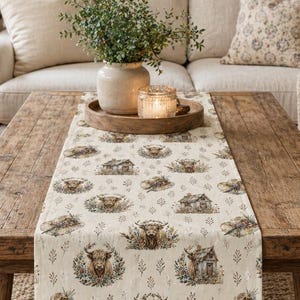 May include: A wooden coffee table with a cream table runner decorated with Highland cows and small cabins. The runner is placed on the table in a living room with a beige sofa and framed artwork. The table is made of wood.