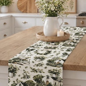 May include: A decorative table runner with a repeating pattern of white cottages, rocking chairs, and floral arrangements in green and white. The runner is displayed on a wooden kitchen island, adding a touch of country charm to the space.