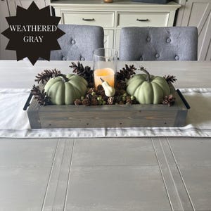 Puede incluir: Una bandeja de madera gris envejecida con asas negras, que contiene dos calabazas verdes, pi&ntilde;as y una vela en un cilindro de vidrio. Tambi&eacute;n se incluye una peque&ntilde;a calabaza blanca. El texto "WEATHERED GRAY" est&aacute; en forma de estrella.