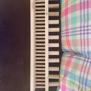 May include: A light-colored wooden wine rack with a ladder-like design. The rack is made of vertical and horizontal wooden slats. The rack is on a brown surface, and a plaid pillow is in the background.