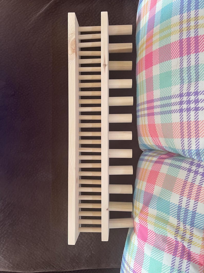May include: A light-colored wooden wine rack with a ladder-like design. The rack is made of vertical and horizontal wooden slats. The rack is on a brown surface, and a plaid pillow is in the background.