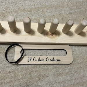 May include: A light-colored wooden rack featuring a row of vertical cylindrical pegs and a separate rectangular wooden tag with rounded edges. The tag has a black elastic loop attached and the text JR Custom Creations engraved in black script.