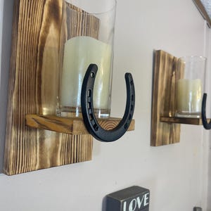 May include: Rustic wall-mounted candle holders. Each features a wooden base with a shelf, a clear glass cylinder, and a black horseshoe accent. The candles are a soft yellow color, creating a warm ambiance.
