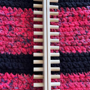 May include: A light-colored wooden drying rack with multiple horizontal dowels. The rack is positioned vertically against a textured background of pink, black, and purple yarn. The rack is made of two vertical pieces connected by the dowels.